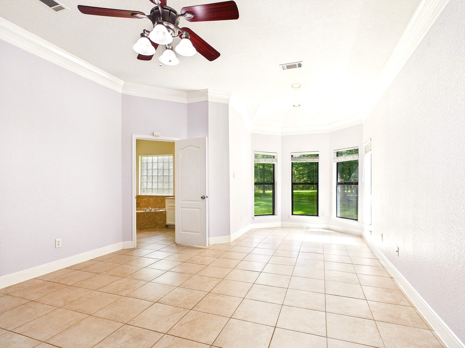 19521 Desna Drive, Porter, TX 77365 This spacious primary bedroom features tiled flooring, a ceiling fan with lights, and ample natural light from large windows with a view of greenery. Adjacent is a bathroom with a corner bathtub.