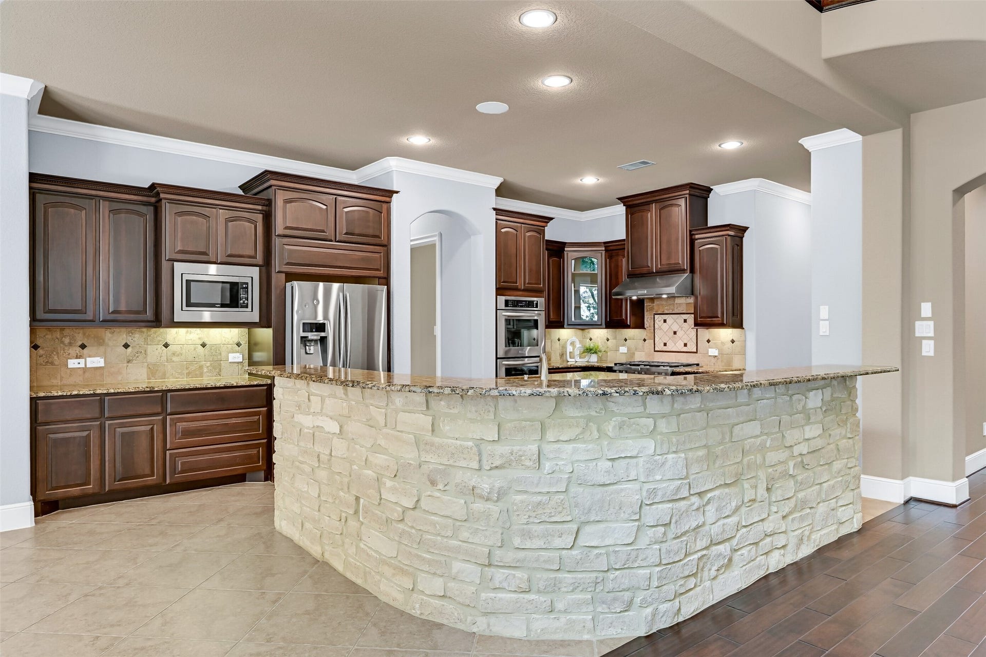 2304 Halls Creek Court, Friendswood, TX 77546 The island kitchen blends warmth and function with rich espresso cabinetry, granite countertops, undercabinet lighting, a travertine tile backsplash, and an oversized wraparound breakfast bar finished with stacked stone detailing that gives the space a custom feel.
