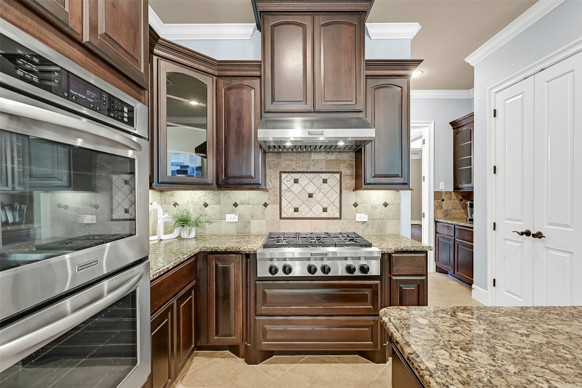 2304 Halls Creek Court, Friendswood, TX 77546 Designed for serious cooking, the kitchen features a six-burner KitchenAid gas cooktop beneath a vent hood framed by custom cabinetry and travertine backsplash details, along with dual electric convection ovens and abundant storage throughout.