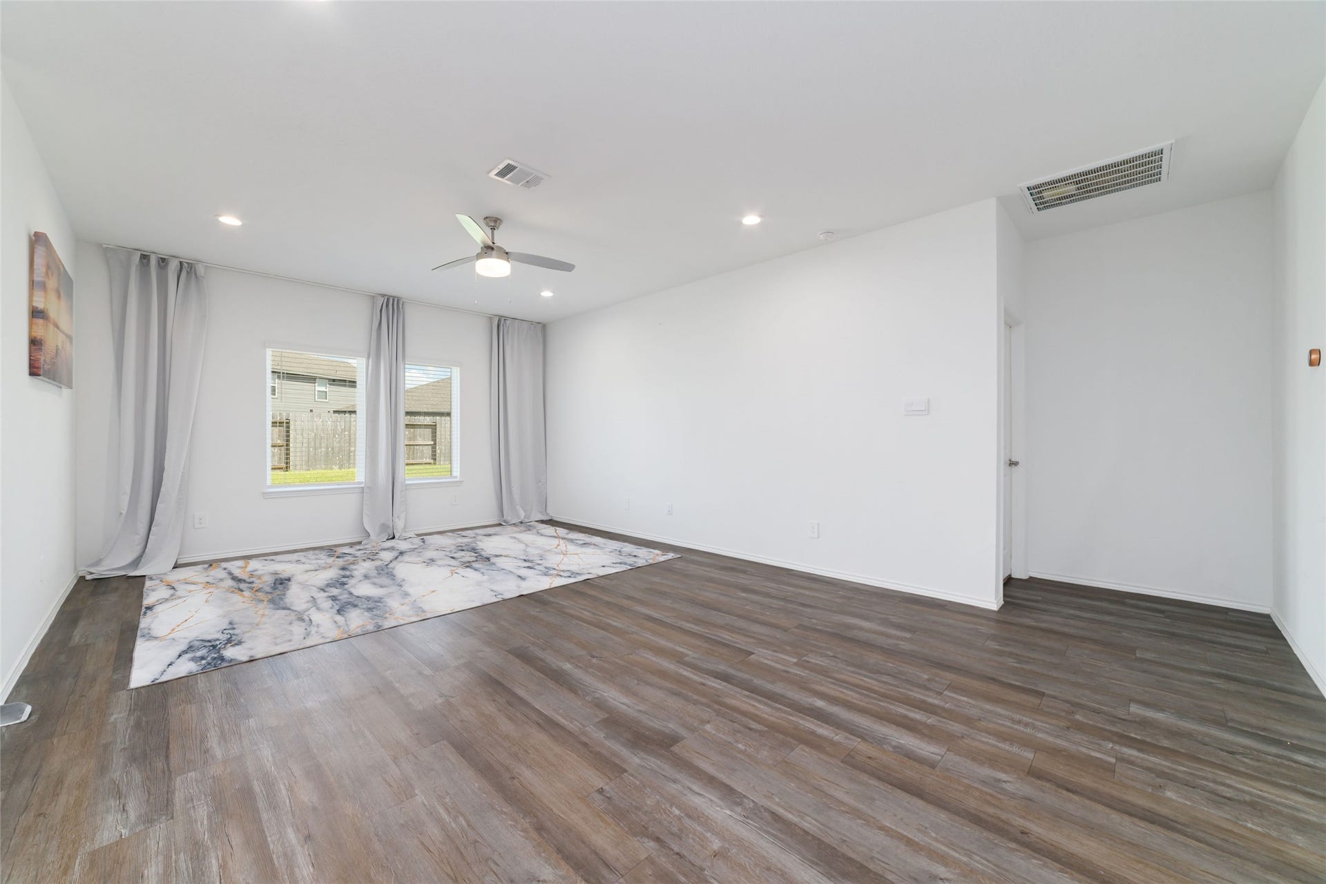 (Main Living Room): Sprawling, light-filled family room designed for effortless entertaining and relaxation. The space boasts continuous dark wood-look flooring, crisp white walls, recessed lighting, and a modern ceiling fan, while twin double windows framed by custom floor-to-ceiling drapery offer peaceful, unobstructed views of the private backyard.