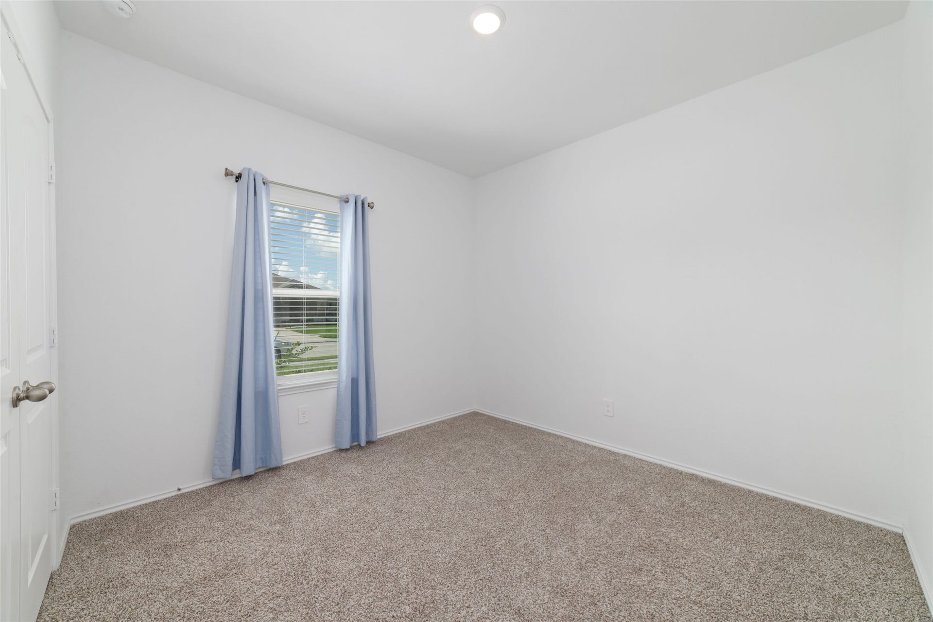 Interior shot of an empty bedroom with beige carpeting, white walls, and a single window dressed with blinds and simple blue curtains, offering a peaceful view outside.
