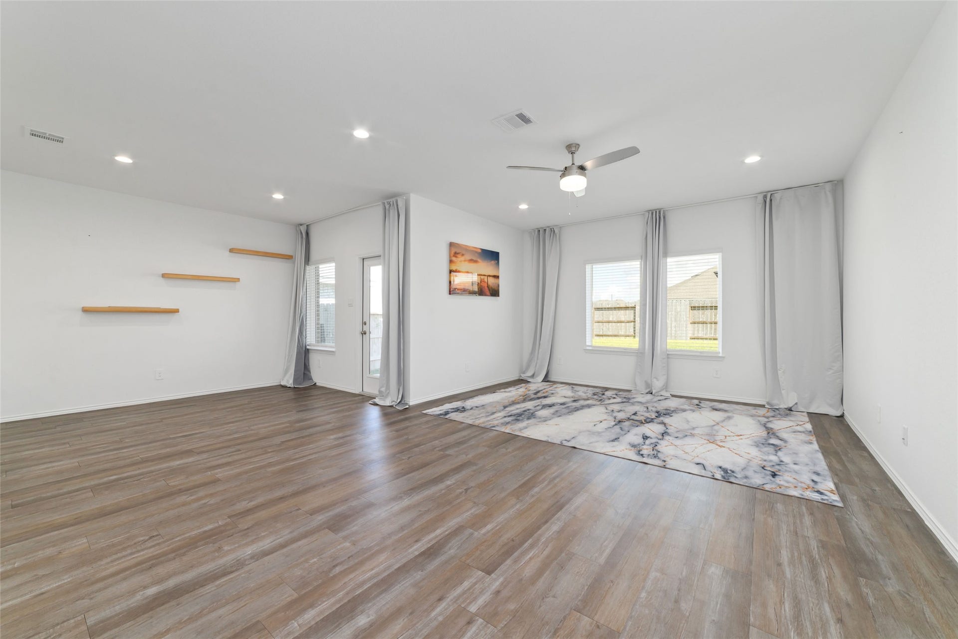 (Main Living Room): Sprawling, light-filled family room designed for effortless entertaining and relaxation. The space boasts continuous dark wood-look flooring, crisp white walls, recessed lighting, and a modern ceiling fan, while twin double windows framed by custom floor-to-ceiling drapery offer peaceful, unobstructed views of the private backyard.