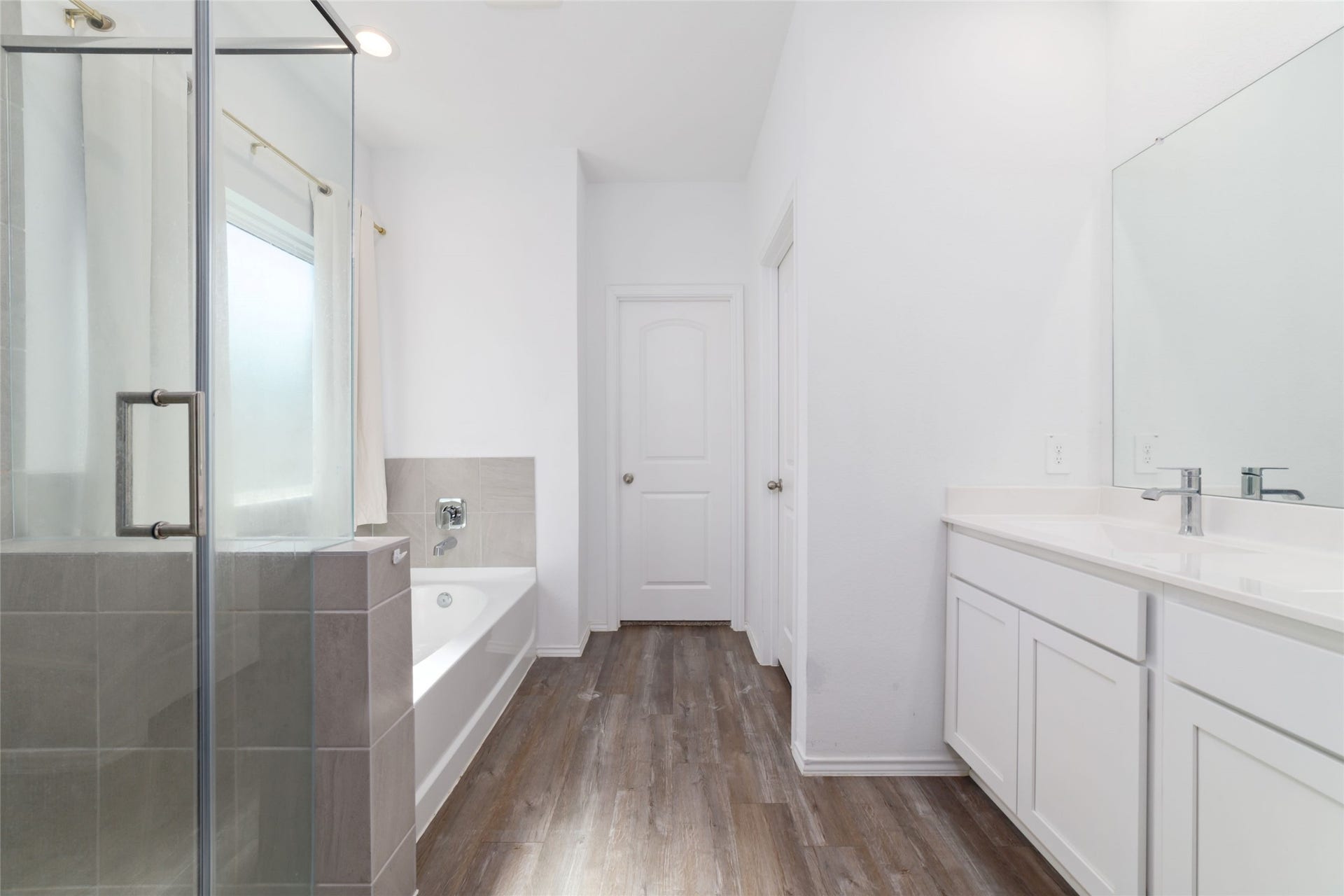 Interior view of a modern full bathroom featuring dark wood-look flooring, a white vanity with an undermount sink, a large mirror, and a tiled shower/bathtub combination with standard fixtures