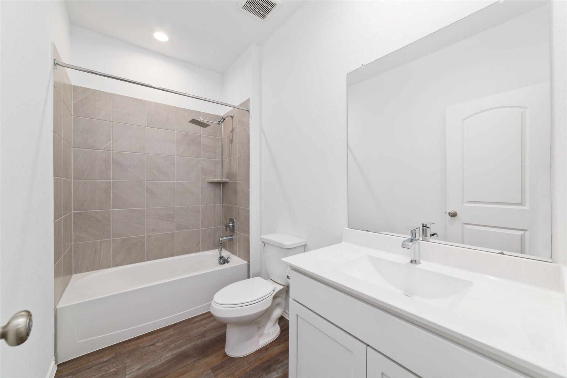 Bright and modern first-floor full bathroom featuring dark wood-look flooring, a sleek white vanity with an undermount sink and expansive mirror, and a tiled shower/bathtub combinationperfectly positioned to service the downstairs secondary bedroom and main-level guests