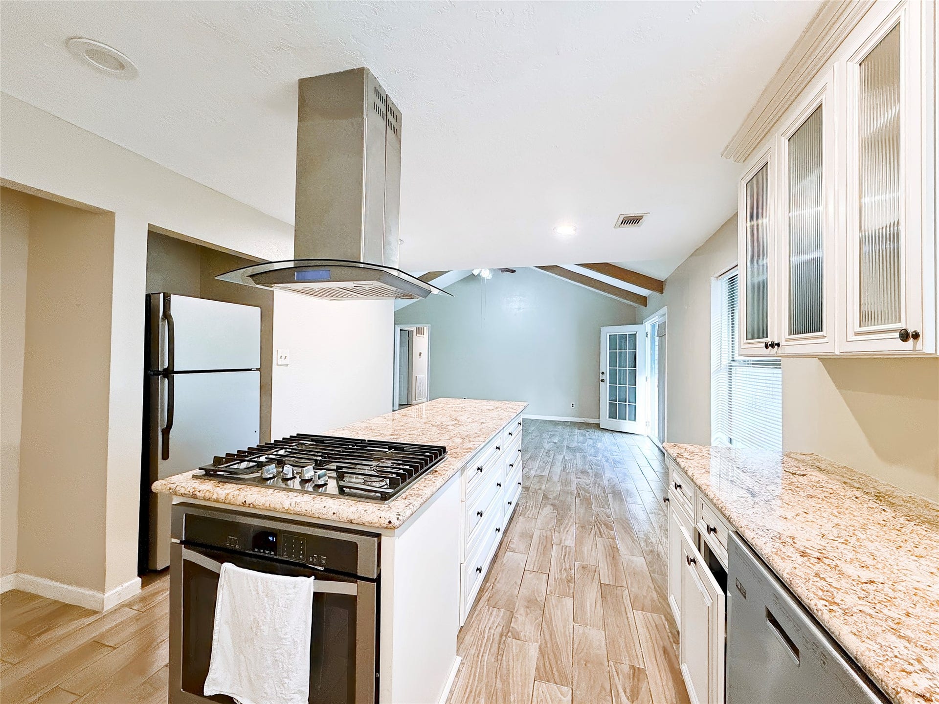 8116 Gulick Lane, Houston, TX 77075 Modern kitchen with granite countertops, a central island featuring a gas cooktop, and stainless steel appliances. The open layout flows seamlessly into a bright living area with vaulted ceilings and wood-look flooring.