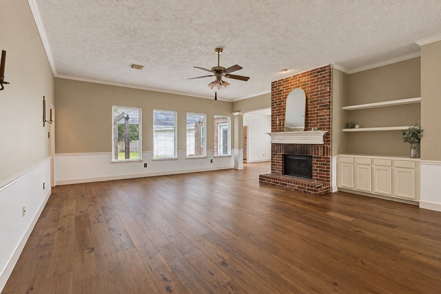 304 Melodywood Drive, Friendswood, TX 77546 This large family room offers generous space for gathering and entertaining, with engineered wood flooring and a brick wood-burning fireplace as its centerpiece. Built-in shelving and cabinetry provide storage and display space, while crown molding and classic wainscoting add character. Four tall windows fill the room with natural light and offer views of the backyard.