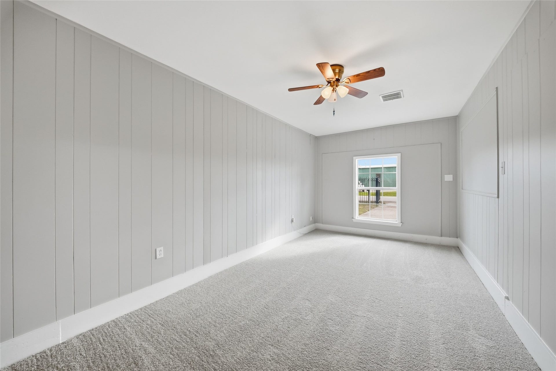 Bright room with soft gray paneled walls, plush carpeting, and a ceiling fan, featuring a large window that invites natural light.