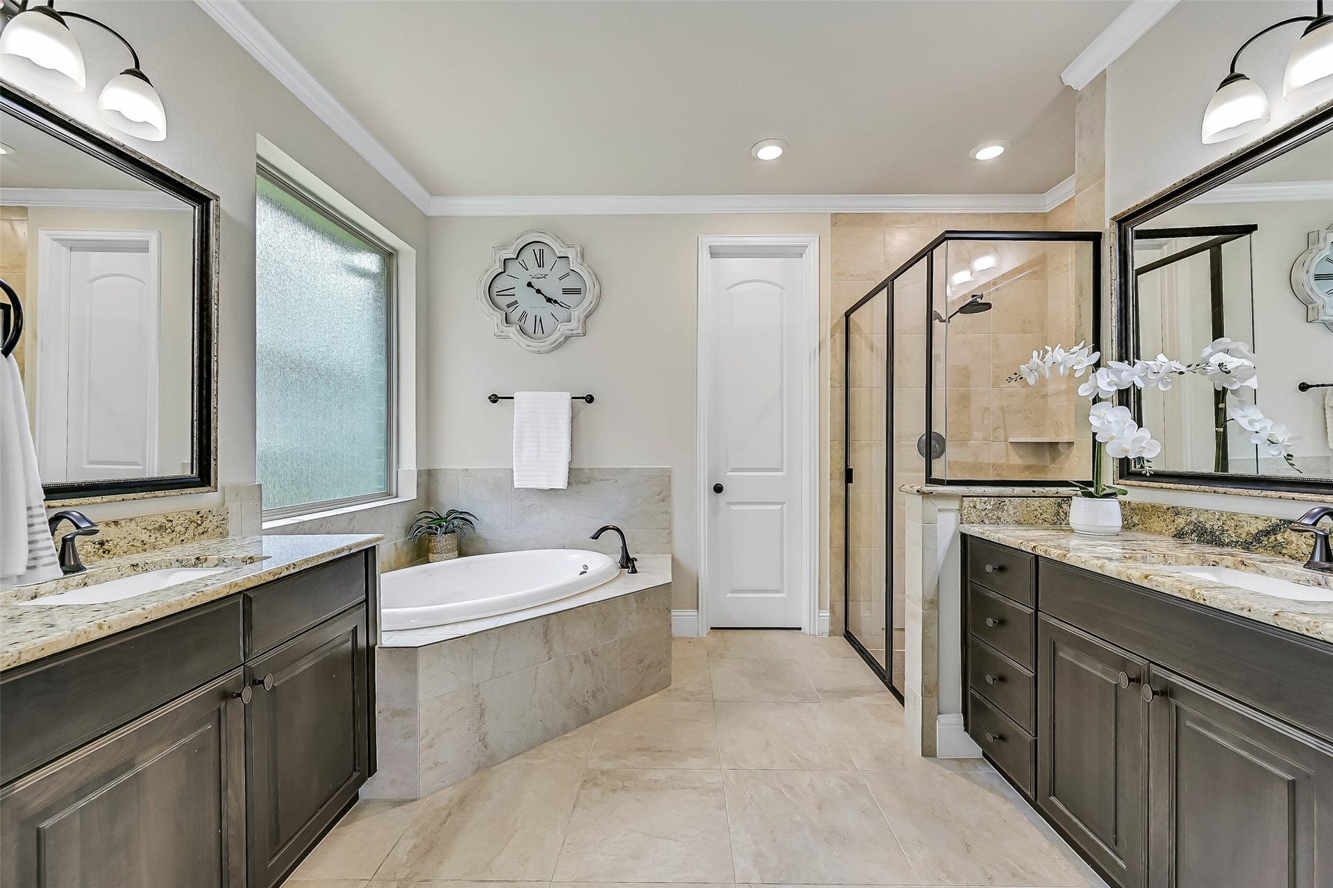 1645 Longwood Glen Lane, Friendswood, TX 77546 The spa-inspired primary bath offers separate granite-topped vanities with framed mirrors, stained wood cabinetry, a relaxing corner soaking tub beneath a privacy glass window, and an oversized glass-enclosed shower with tile surround and built-in shaving ledge.