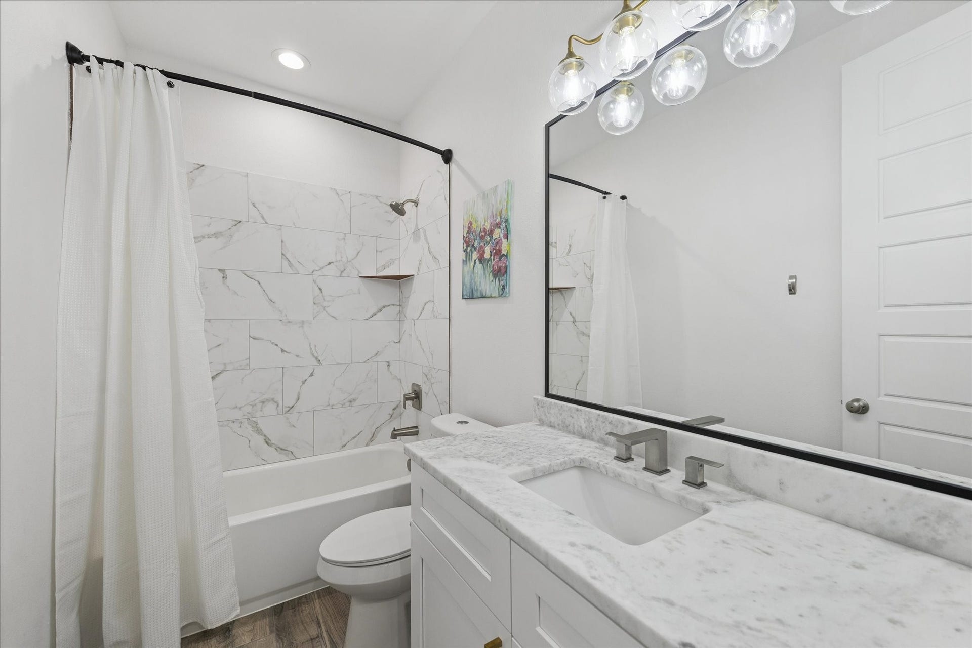 Every detail in this home reflects high-end quality, as seen in this secondary bathroom. The marble countertops, clear tile in the bathtub area, and custom mirror with dark edging create a harmonious and sophisticated look. Recently installed dark faucets and the brass LED light fixture enhance the space, making it both functional and stylish. This serene bathroom offers a calm and inviting retreat for family members or guests.
