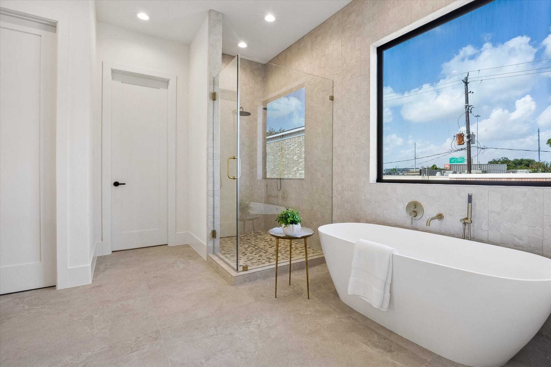 335 Harvard Street, Houston, TX 77007 Freestanding soaking tub sits beneath a large picture window, while the adjacent walk in shower features floor to ceiling stone tile, dual shower heads, and warm brass fixtures.