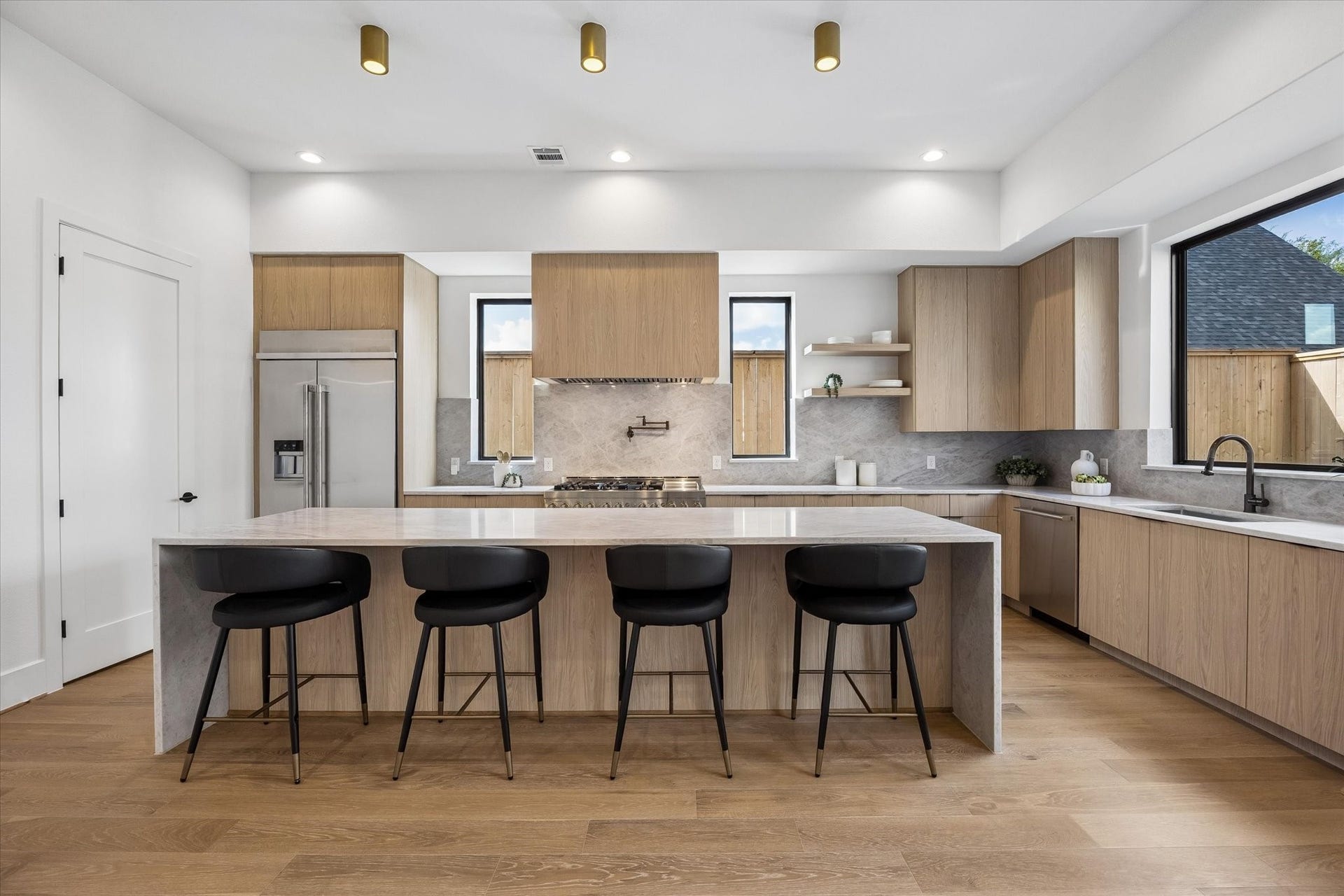 335 Harvard Street, Houston, TX 77007 Natural oak cabinetry gives the kitchen a warm, tailored look, complemented by a stone backsplash that carries seamlessly behind the range. An expansive waterfall island offers seating for four, while slim windows and curated open shelving keep the design light and architectural.