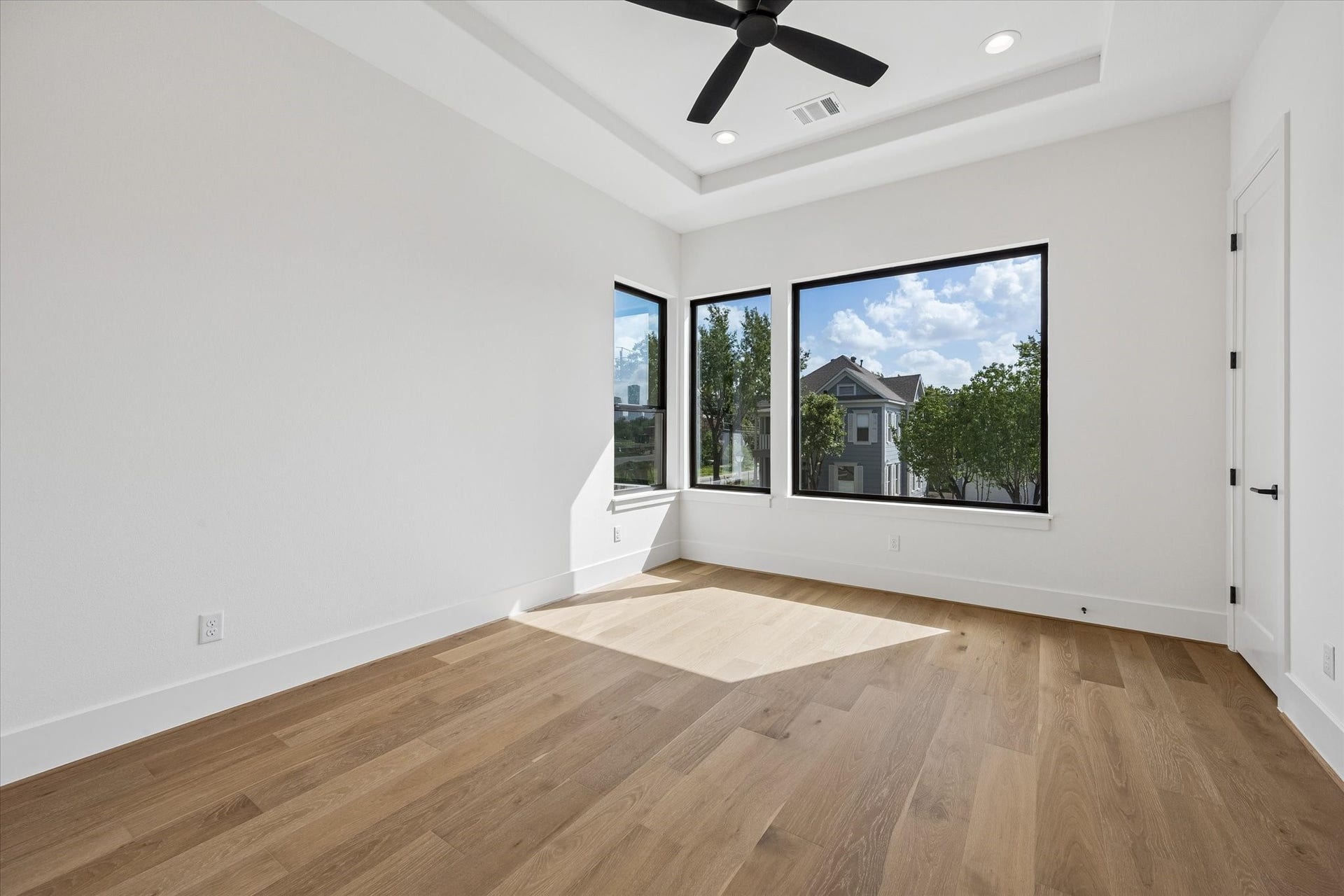 335 Harvard Street, Houston, TX 77007 Natural light pours into the secondary bedroom through oversized black framed windows, complemented by warm hardwood floors and a tray ceiling that adds architectural dimension.