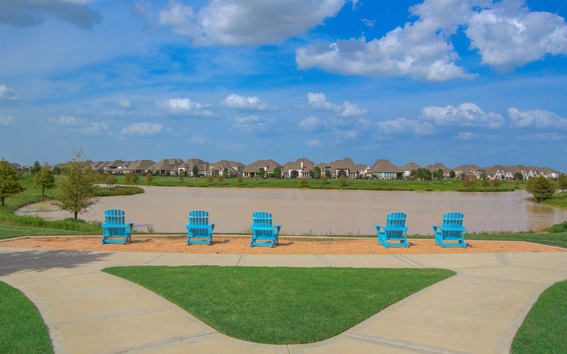 24430 Switchgrass Valley Way, Katy, TX 77493 Experience ultimate relaxation with this serene lakeside view, featuring a row of inviting blue Adirondack chairs perfectly positioned to overlook the calm water and picturesque neighborhood homes under a bright blue sky.