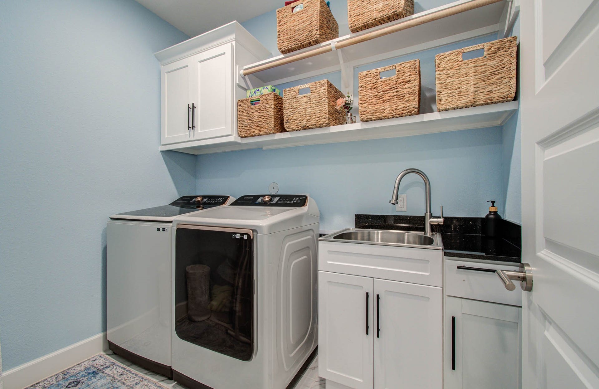 24430 Switchgrass Valley Way, Katy, TX 77493 This bright and functional laundry room features modern appliances, ample storage with overhead cabinets and woven baskets, a convenient sink, and a sleek black countertop, all set against a soft blue wall.