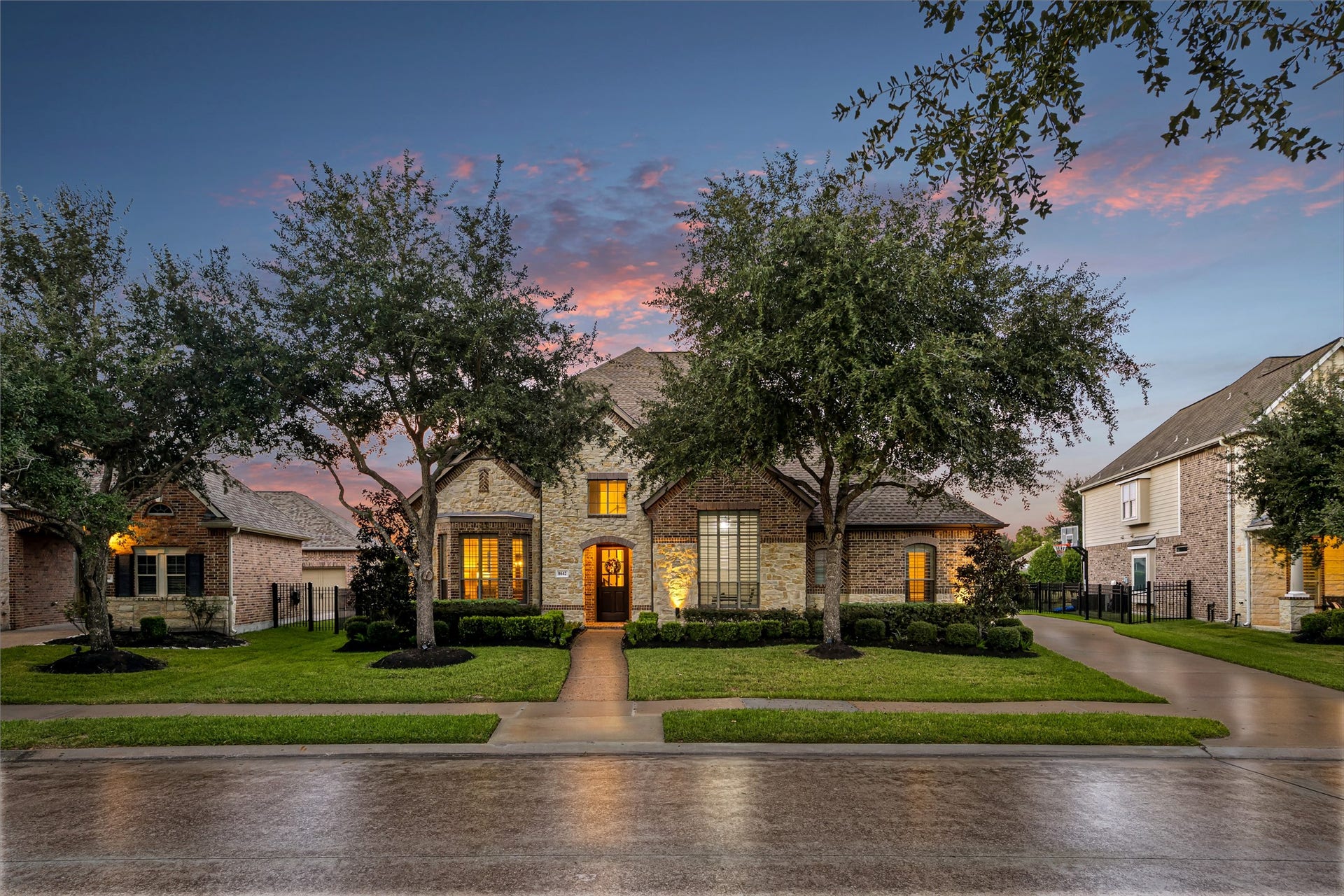 8642 Chickasaw Plum Way, Katy, TX 77494 Stately brick and stone facade glowing at twilight, framed by towering mature oaks and manicured landscaping. New windows (2024). (Virtually enhanced: driveway reflection)