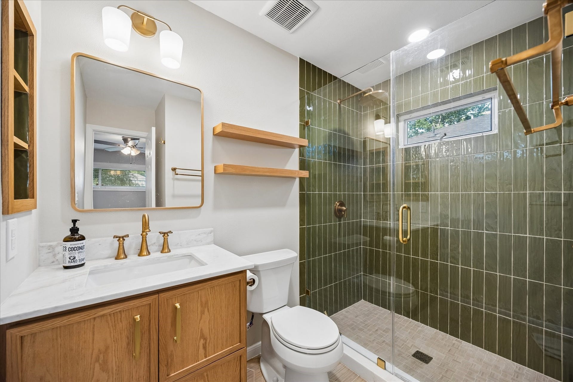 2214 Saxon Drive, Houston, TX 77018 Rich green tile, warm wood cabinetry, and brushed brass fixtures create a striking designer aesthetic in the primary bath. A frameless glass shower, floating wood shelves, and marble look countertops blend timeless finishes with modern style.