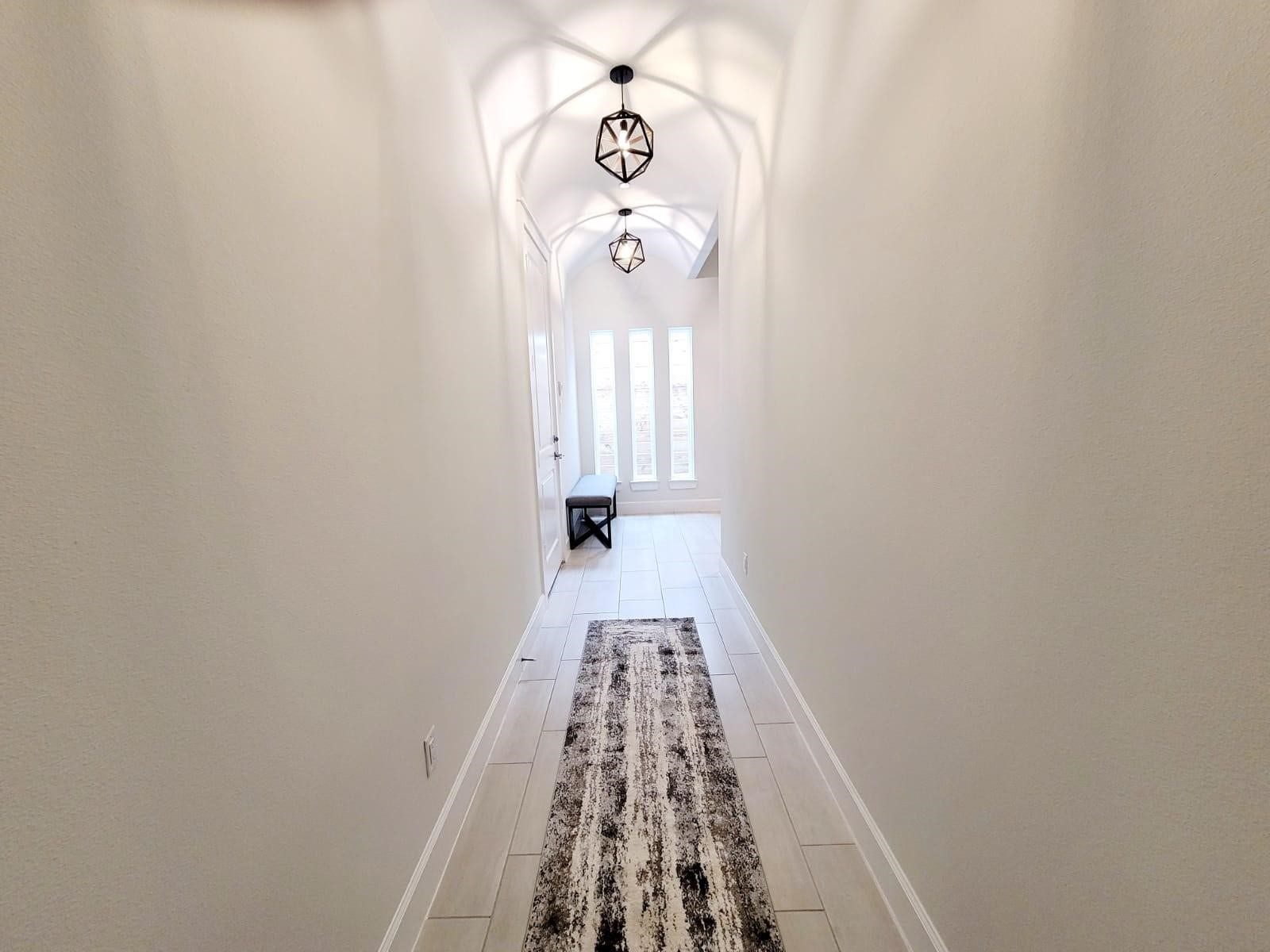 The entry way has a bright, modern hallway with a high ceiling and stylish pendant lights. The space is enhanced by a long rug and natural light streaming through tall windows at the end, creating an inviting entryway.