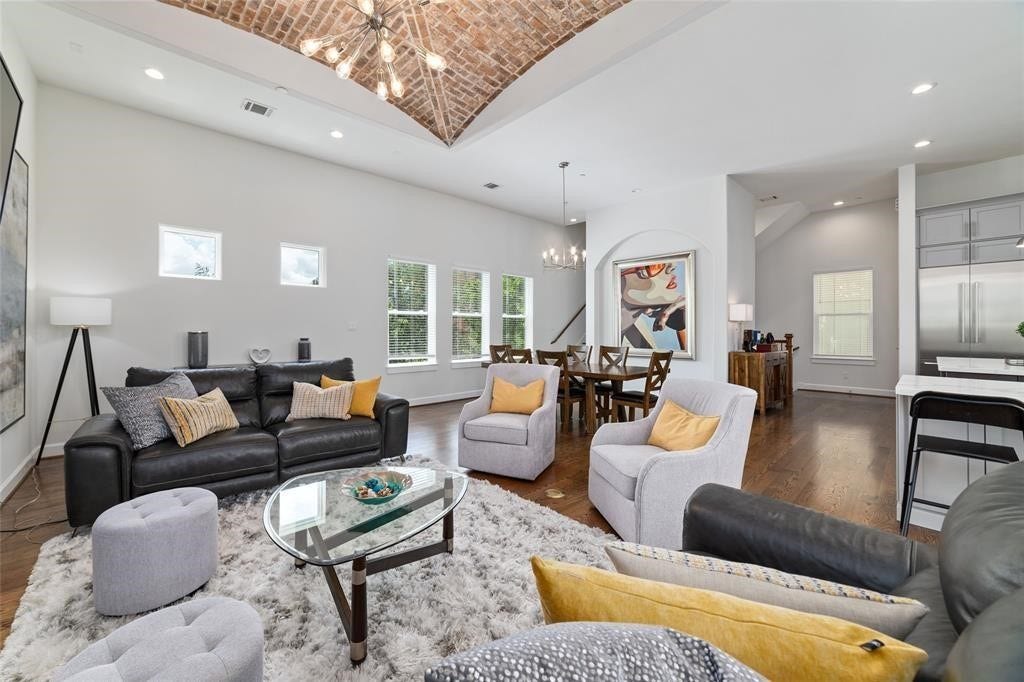 The spacious living area features a modern open-concept design with high ceilings and a stylish brick accent. The room is filled with natural light from multiple windows and includes a cozy seating area, elegant dining space, and a sleek kitchen with stainless steel appliances. Perfect for entertaining and family gatherings.