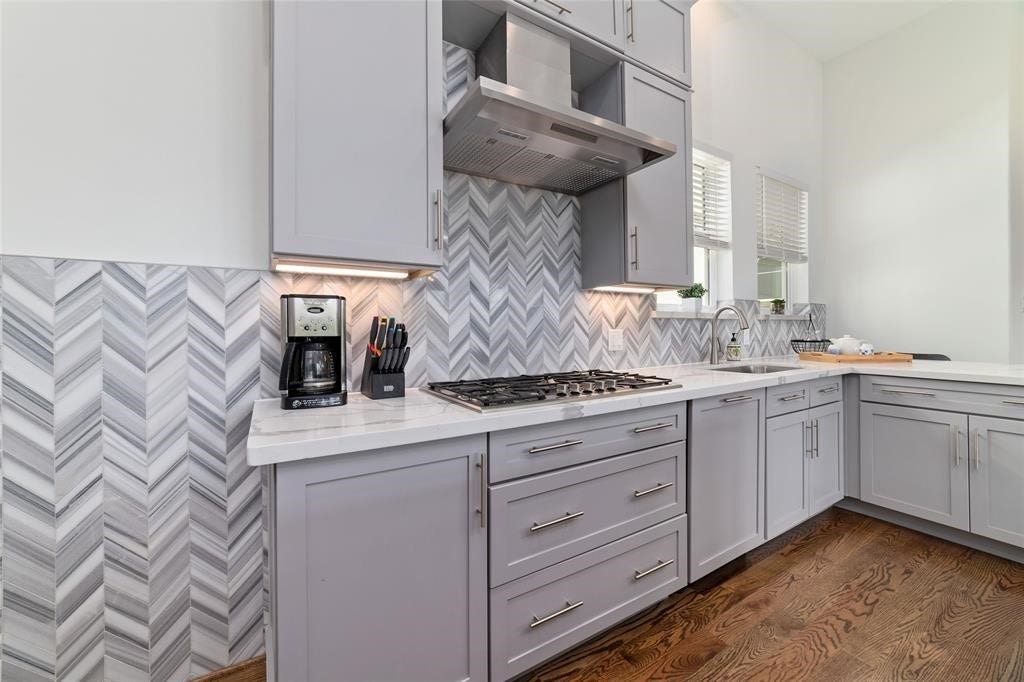 The modern kitchen features sleek gray cabinetry and a stylish chevron-patterned backsplash. It includes a gas stove, stainless steel range hood, and ample counter space with a built-in sink, all set against warm hardwood floors.