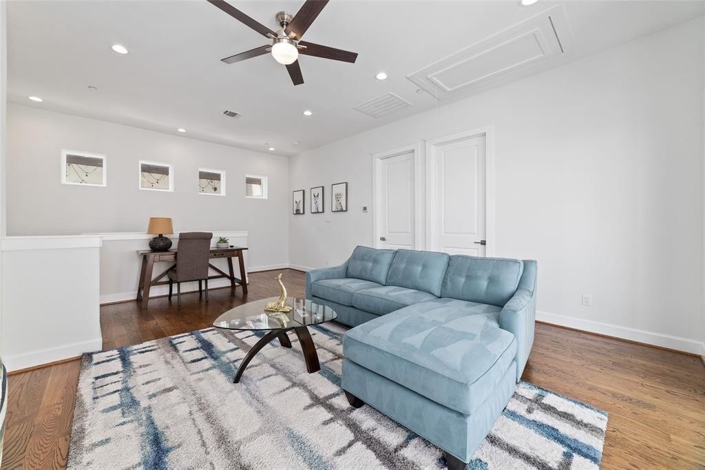 This bright, modern living space features a cozy blue sectional sofa, a stylish glass coffee table, and a sleek ceiling fan. The room is well-lit with recessed lighting and offers a small workspace with a desk and chair, all set on beautiful hardwood floors.