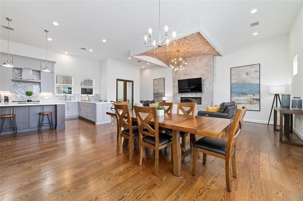 This spacious open-concept living area features a modern kitchen with a large island, stylish herringbone backsplash, and pendant lighting. The dining area flows seamlessly into the cozy living room, which boasts a stone fireplace and contemporary decor. High ceilings and hardwood floors enhance the airy feel.