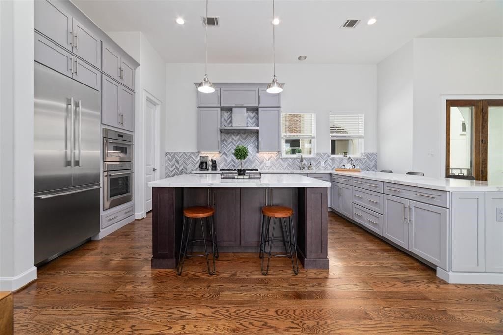 The kitchen features modern gray cabinetry, a spacious quartz island with seating, stainless steel appliances, and hardwood floors. The backsplash has a stylish herringbone pattern, and pendant lighting adds a contemporary touch.