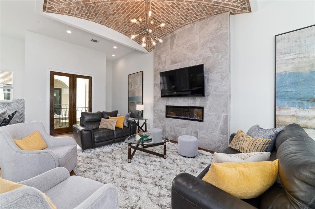 This inviting living room features a vaulted brick ceiling, a modern stone accent wall with a built-in fireplace and TV, cozy seating with stylish cushions, and natural light from double doors. A plush carpet completes the warm, elegant aesthetic.