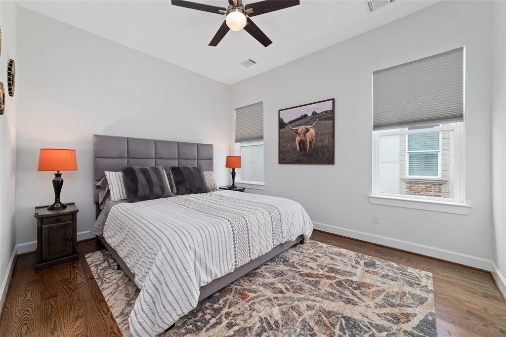 This bedroom features a modern design with hardwood floors, a plush bed, and stylish decor. It includes two windows with shades, a ceiling fan, and warm lighting from bedside lamps, creating a cozy and inviting atmosphere.