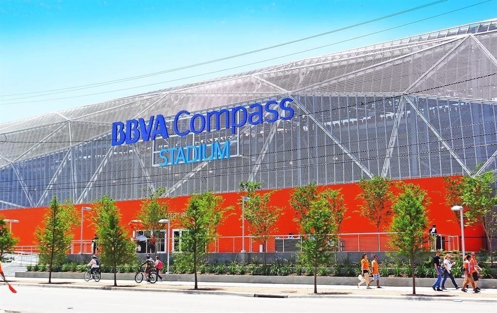 The exterior of the soccer stadium with a distinctive geometric design and vibrant orange accents. The area is pedestrian-friendly, with people walking and biking nearby, and is bordered by young trees, a lively community environment.