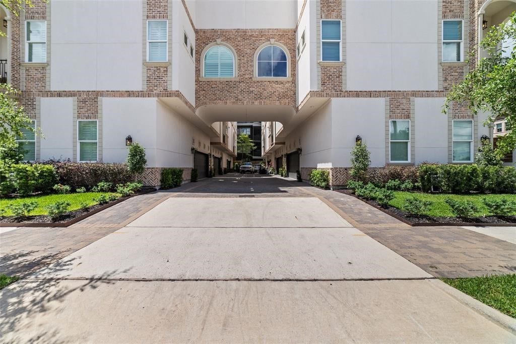 The modern townhouse complex with a shared driveway leading to garages. The building features a mix of brick and stucco exterior, with well-maintained landscaping and greenery on both sides. The architecture includes large windows, and there's a central archway providing access to the inner areas.