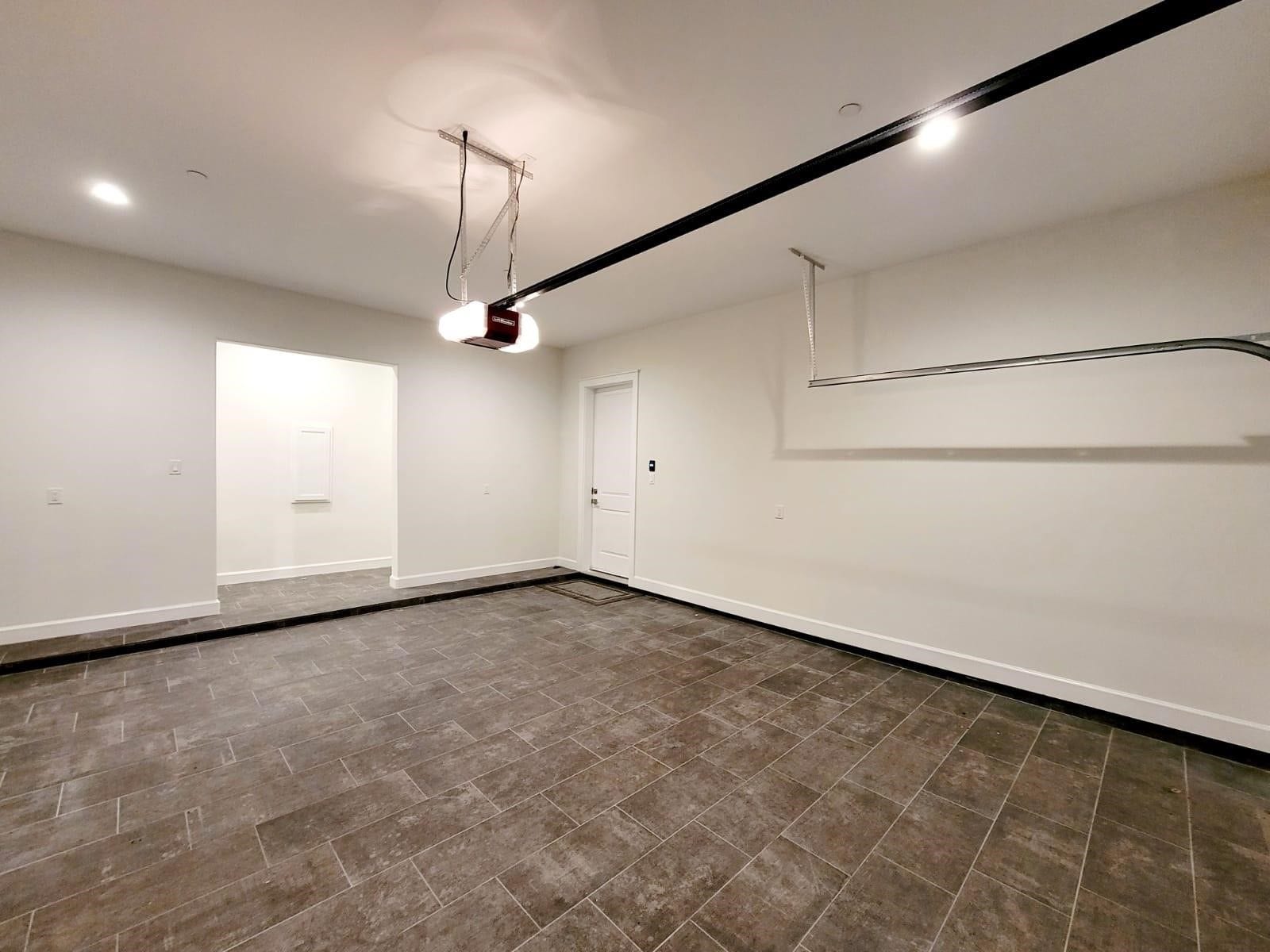 The spacious, clean garage with tiled flooring, white walls, and ceiling lighting. It features a standard garage door opener and an internal door leading into the home.