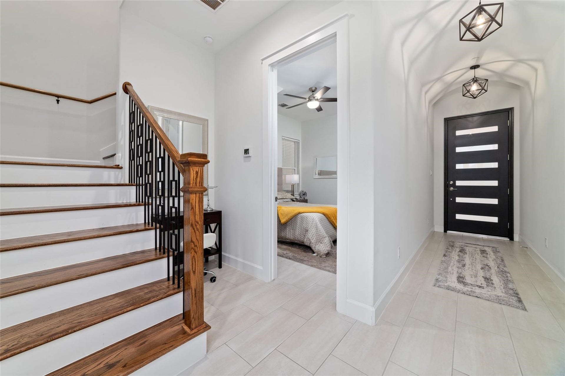 This modern entryway with sleek tile flooring and contemporary lighting fixtures. A stylish staircase with wooden steps and a black railing leads to the upper level. The hallway opens to a room on the right, showcasing a cozy bedroom with a ceiling fan. The front door has a chic, horizontal panel design, adding to the home's modern aesthetic.