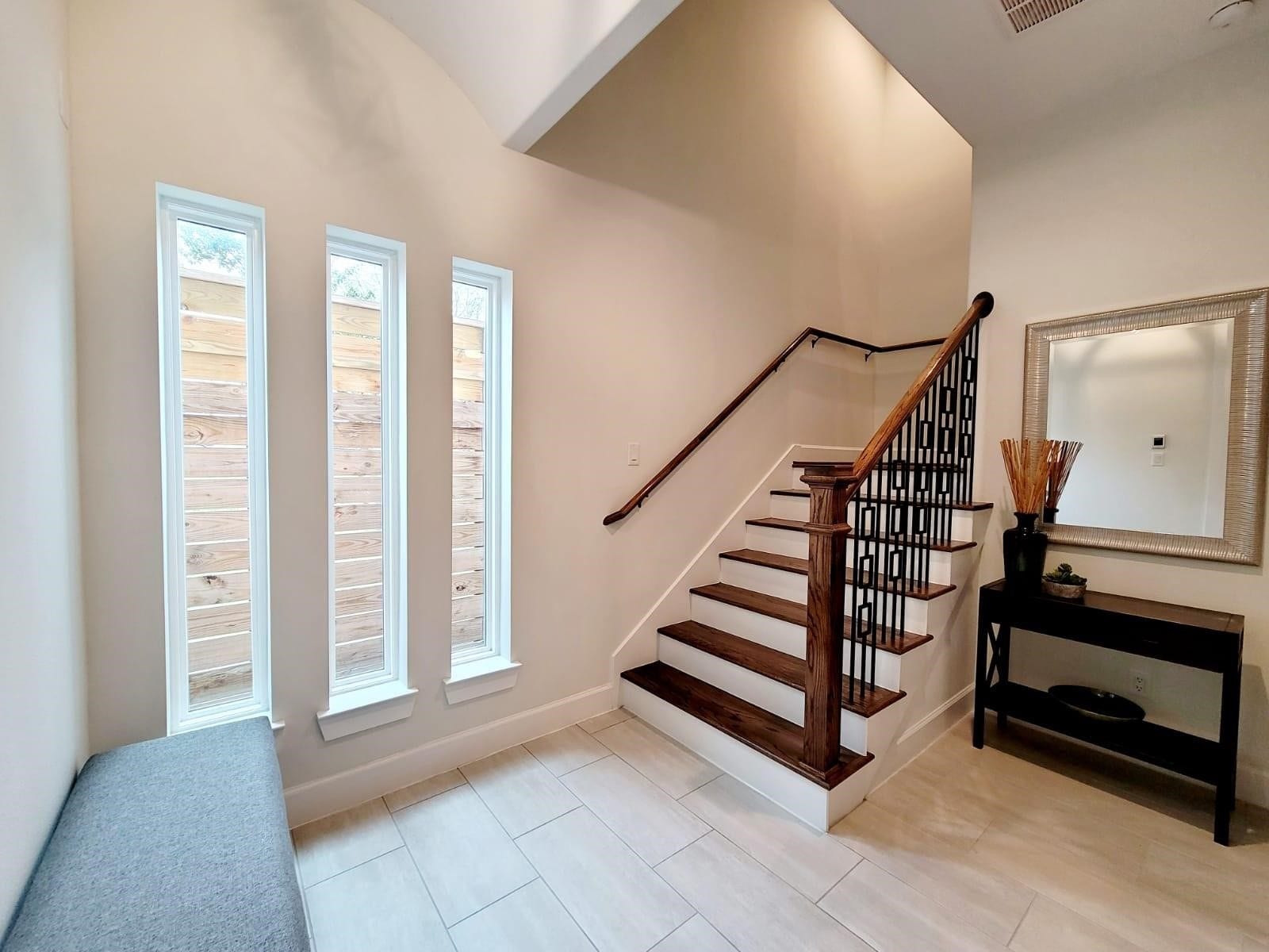 Bright entryway with modern staircase featuring wooden steps and stylish railing. The space includes tall, narrow windows for natural light. Neutral tile flooring complements the clean, contemporary design.