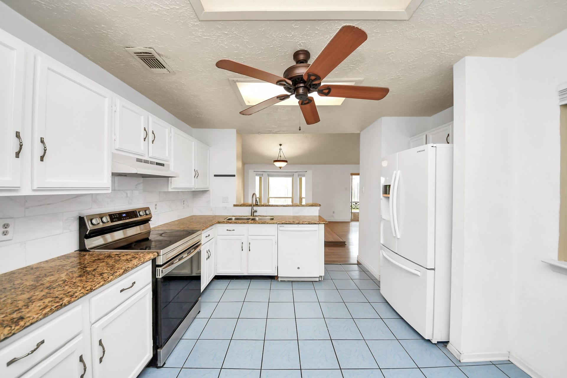 This kitchen features white cabinetry, granite countertops, and a modern stove. It has blue tile flooring and a large ceiling fan. The space opens into a dining area with ample natural light.