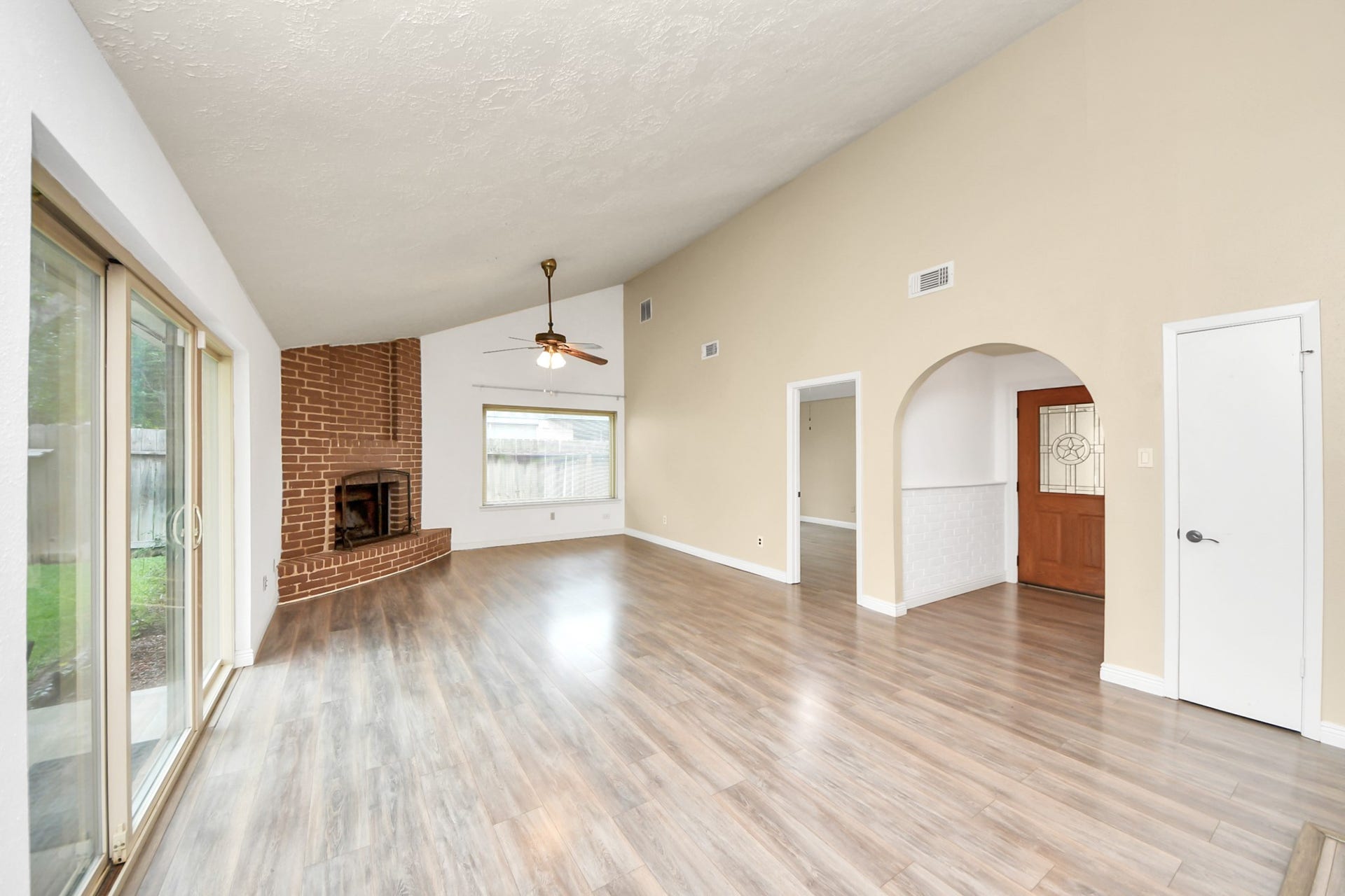 This spacious living room features high ceilings, a brick fireplace, and laminate wood flooring. Large sliding glass doors provide ample natural light, leading to an outdoor area. The neutral walls and open archways create an inviting and versatile space.