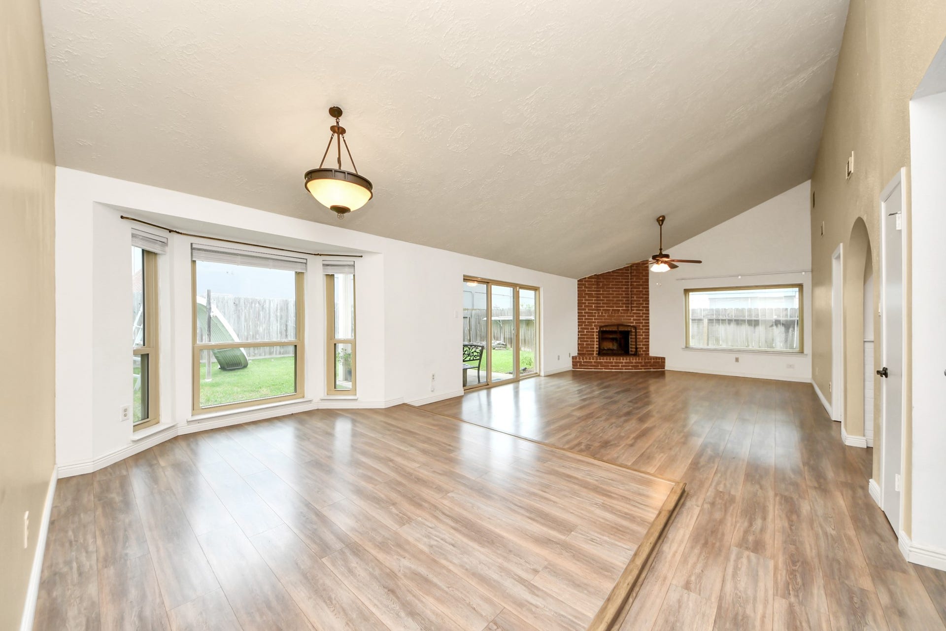 This spacious living area features a vaulted ceiling, abundant natural light from large windows, and elegant wood flooring. It includes a cozy brick fireplace, perfect for gatherings.