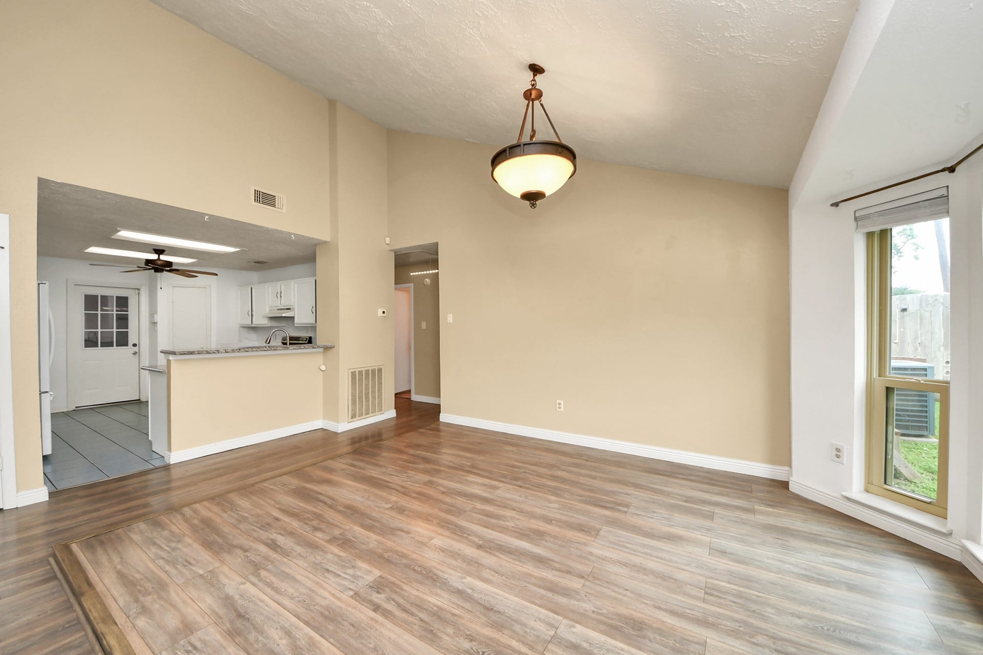This room features an open floor plan with wood-style flooring and neutral walls. It includes a dining area with a hanging light fixture, a large window with natural light, and a view into a modern kitchen with white cabinets and granite countertops.