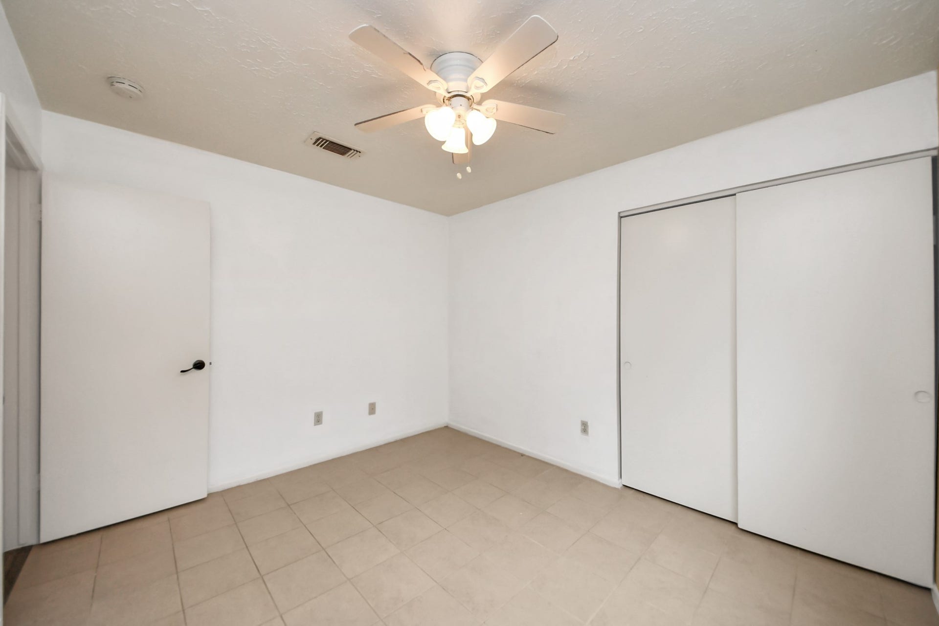 A bright, minimalist room with neutral tile flooring and white walls. It features a ceiling fan with lights and sliding closet doors, offering a clean and functional space.