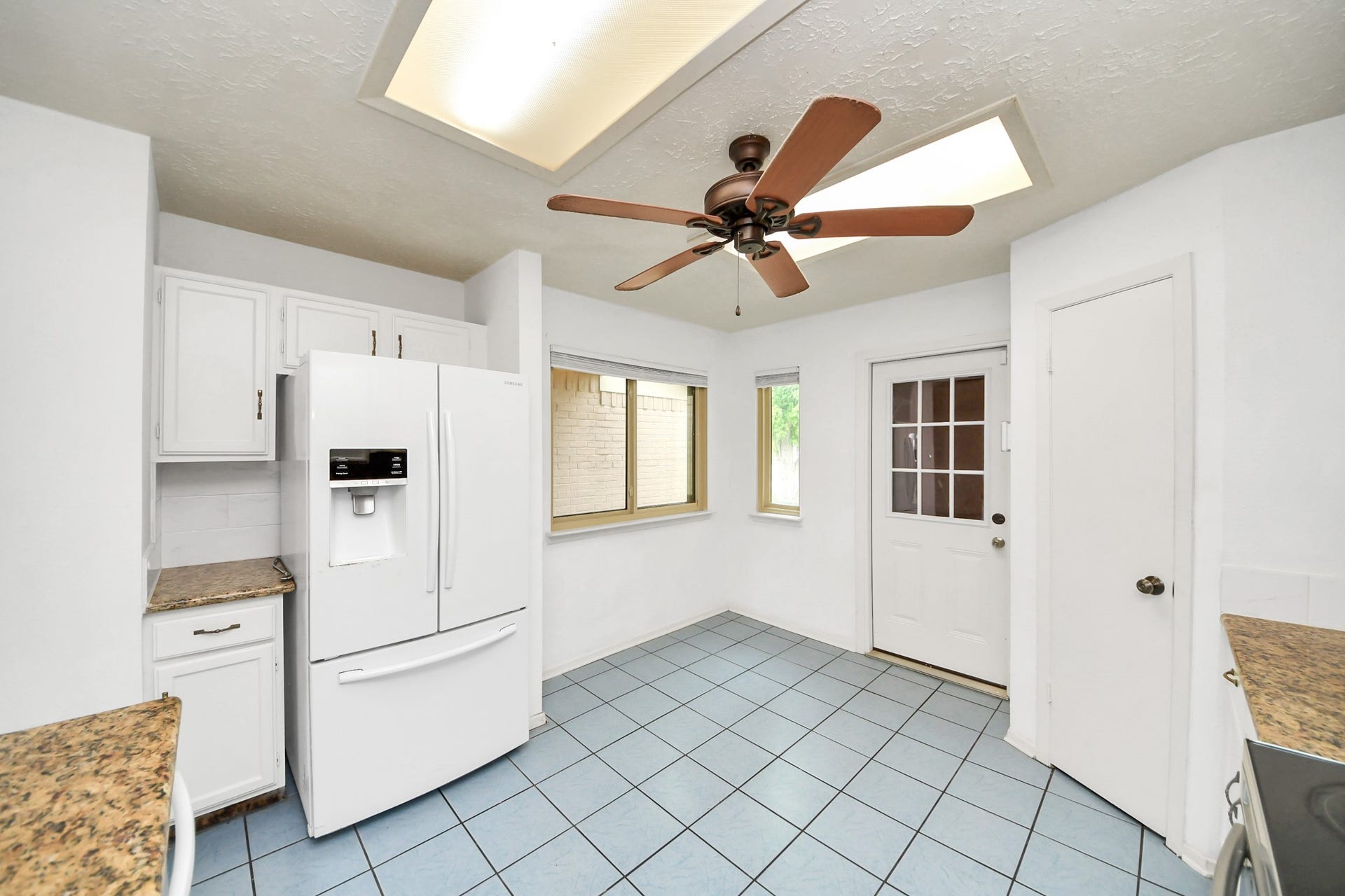 Bright kitchen with white cabinets, granite countertops, and a large refrigerator. Features blue tile flooring, a ceiling fan, and ample natural light from a window and a door with glass panels.