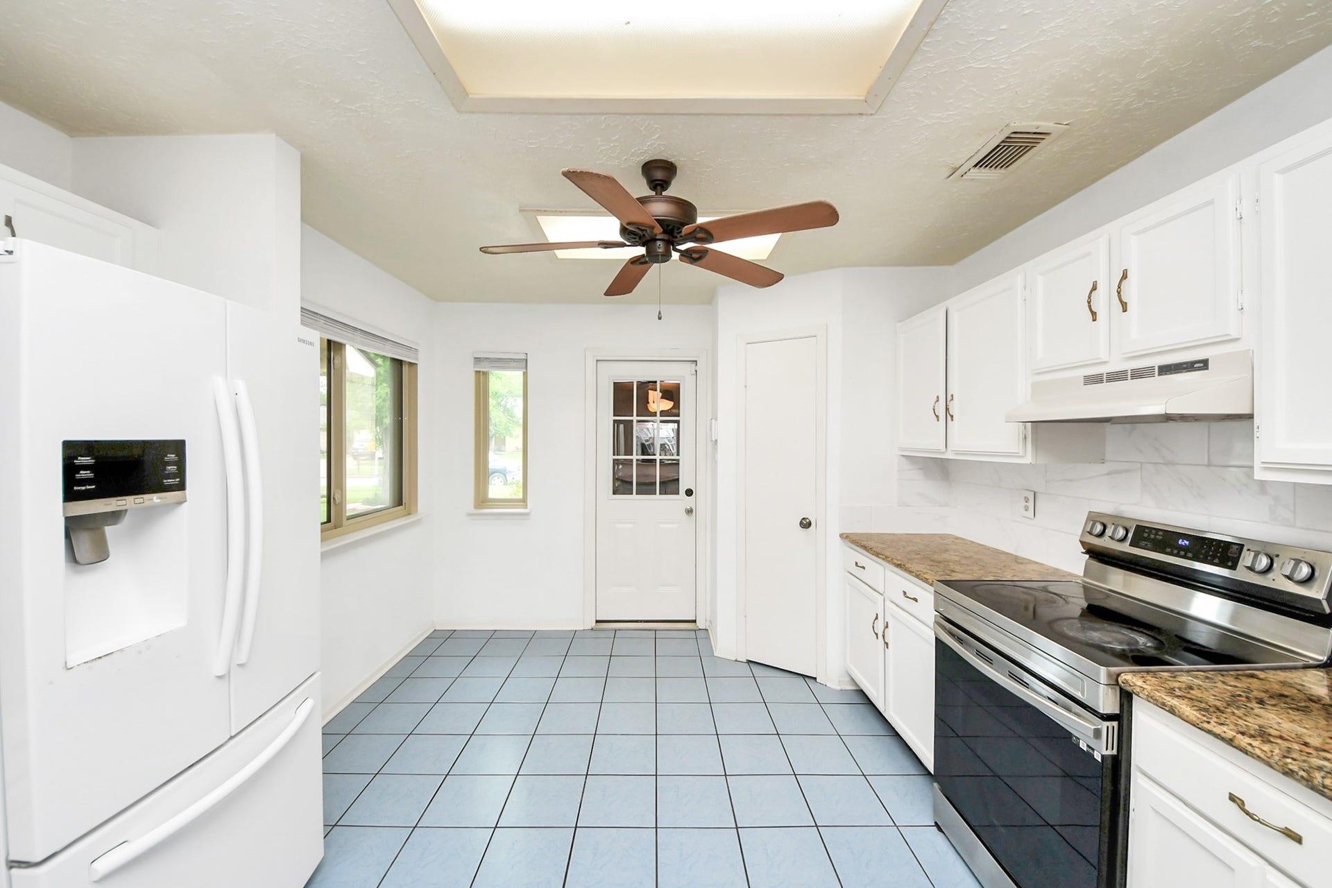 Bright kitchen with white cabinetry, granite countertops, and stainless steel appliances. Features a ceiling fan, tiled floor, and a door leading outside.