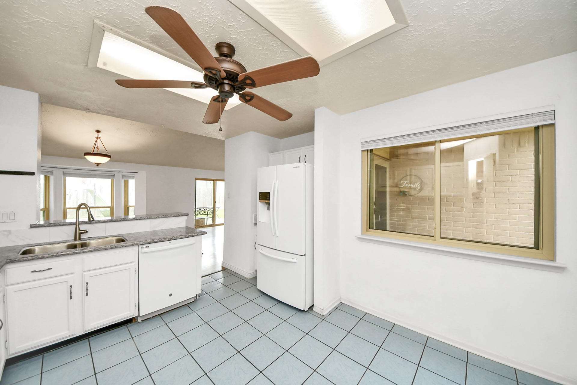 This kitchen features a spacious layout with white cabinetry and appliances, a ceiling fan, and large windows for natural light. It has tile flooring and an adjacent dining area, creating a bright and open space.