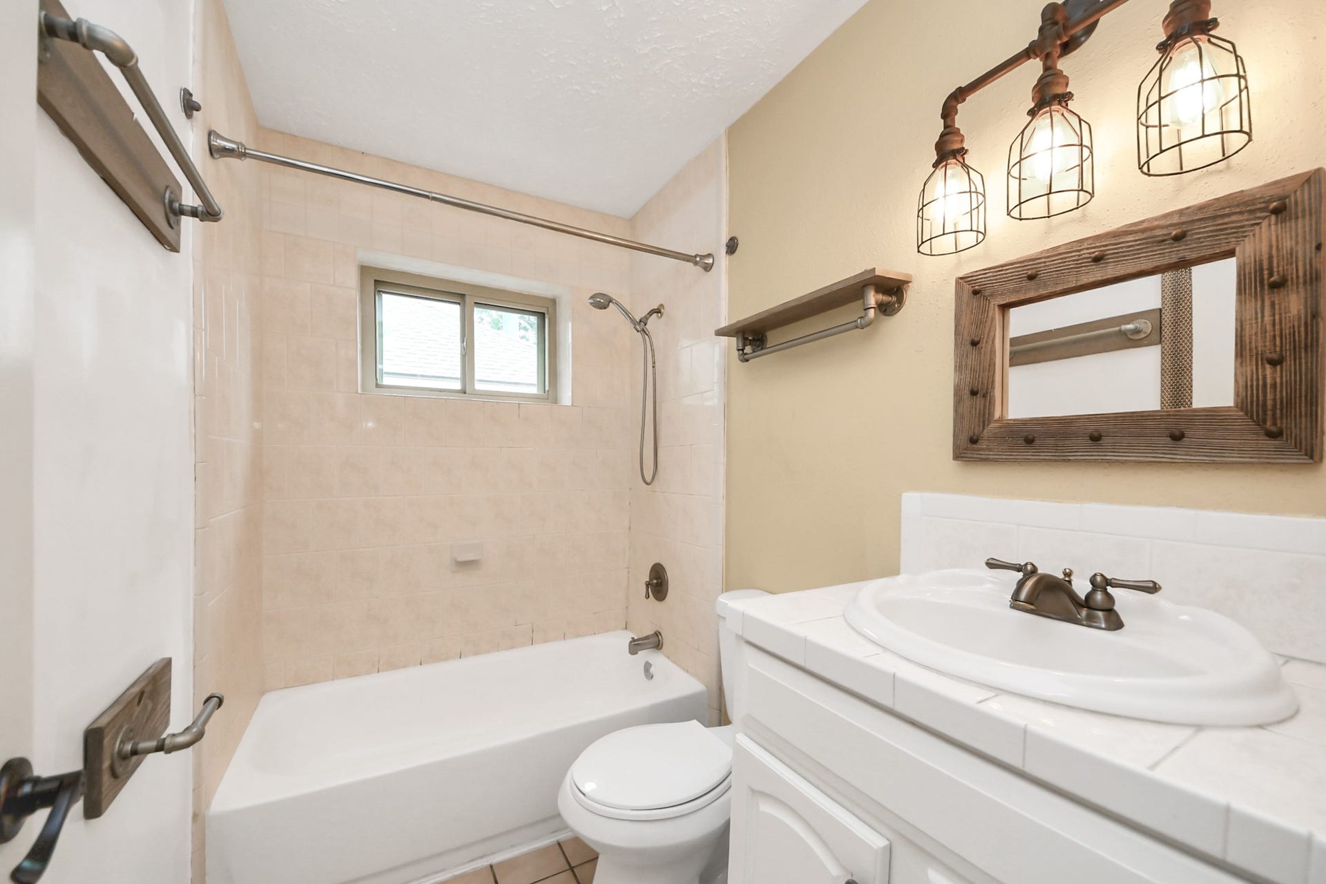 Second bathroom features a cozy layout with a tub-shower combo, a small window for natural light, and rustic fixtures including a framed mirror and industrial-style lighting above the sink.