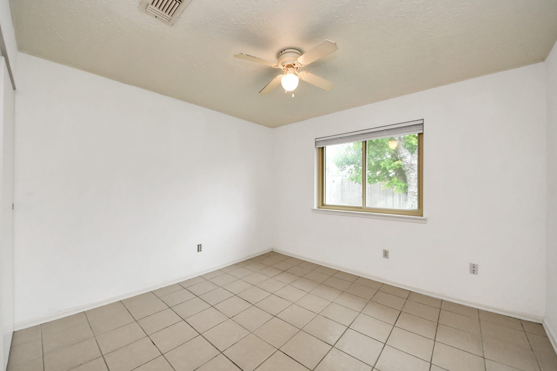 This room features neutral tiled flooring, a ceiling fan, and a large window for natural light. It has white walls and a simple, clean design, ideal for a bedroom or office.