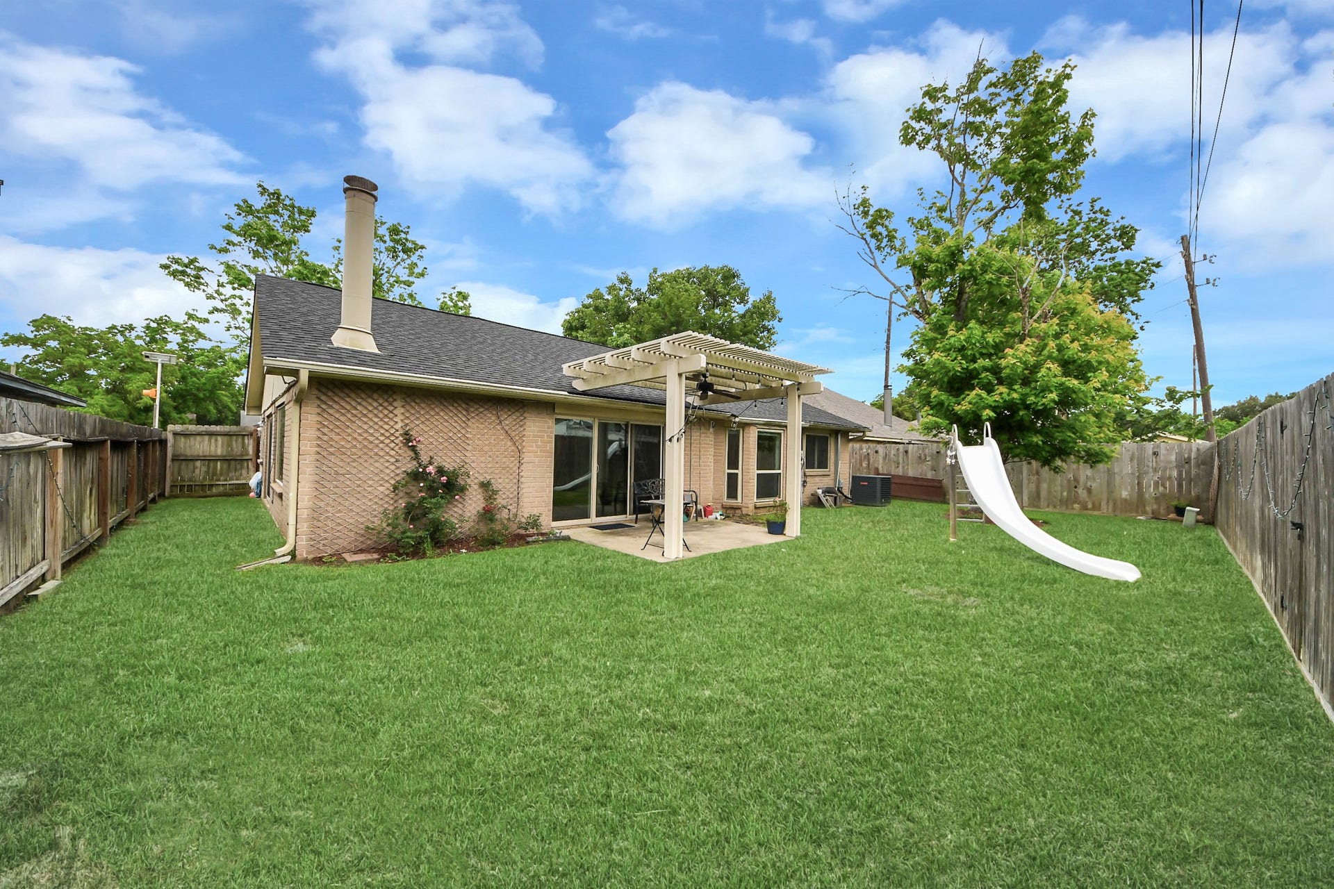 This photo showcases a spacious backyard with a well-maintained lawn, a wooden pergola over a patio area, and a slide. The yard is fenced, providing privacy, and includes mature trees and plants, offering a tranquil outdoor space.