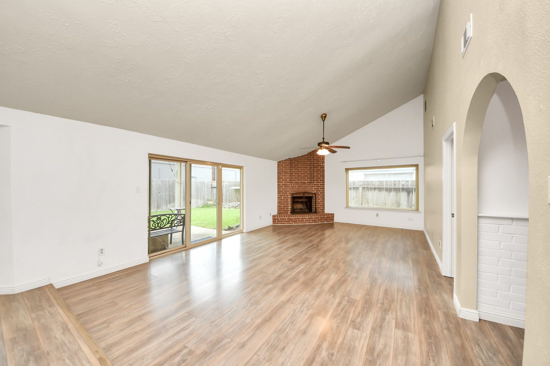 This spacious living room features a high ceiling, laminate wood flooring, and a cozy brick fireplace. Large windows and glass doors provide natural light and lead to a backyard, enhancing the open, airy feel.