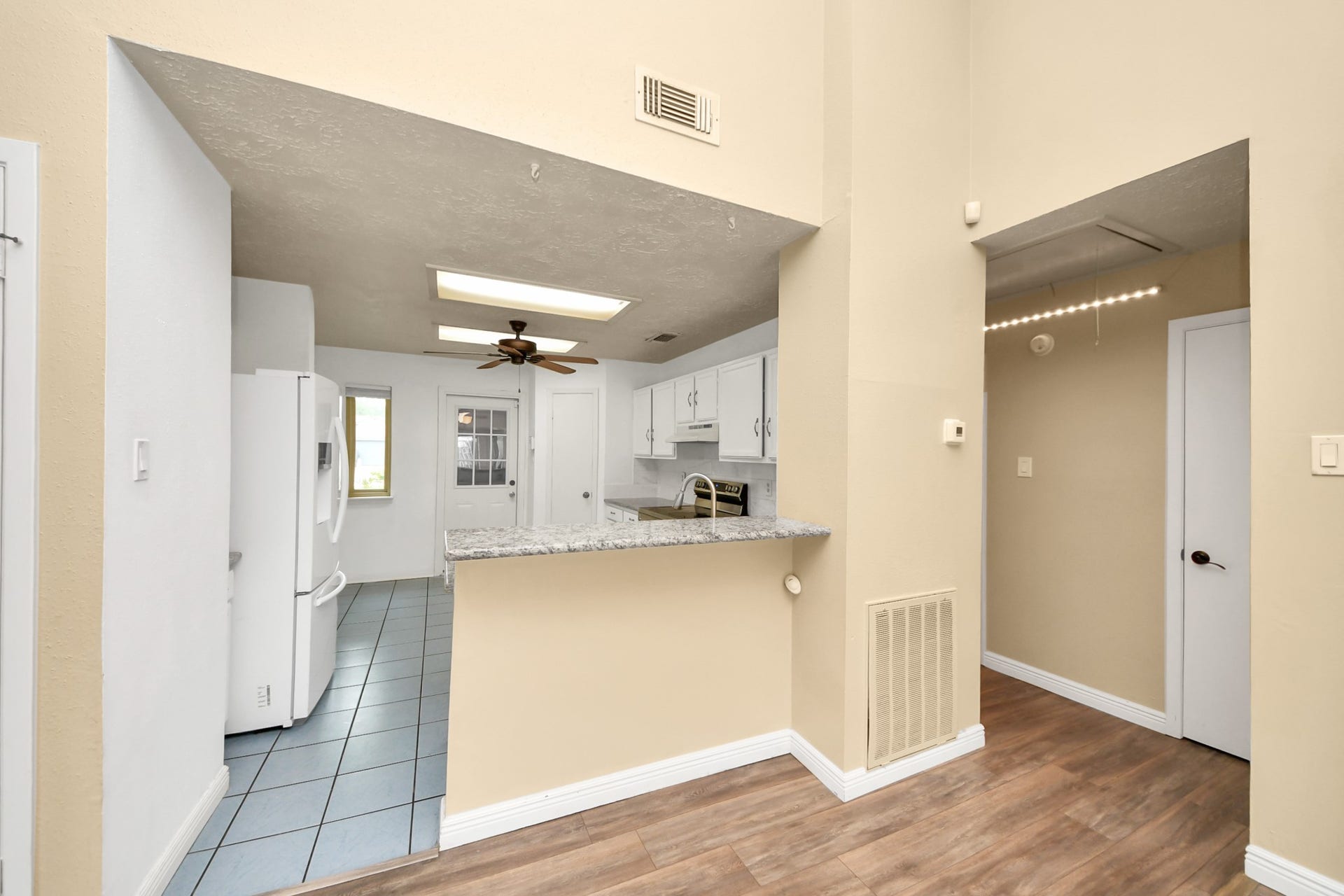 This photo showcases a bright, modern kitchen with granite countertops, white cabinets, and a ceiling fan. The space features tile flooring, a breakfast bar, and transitions to a hallway with wood-style flooring.