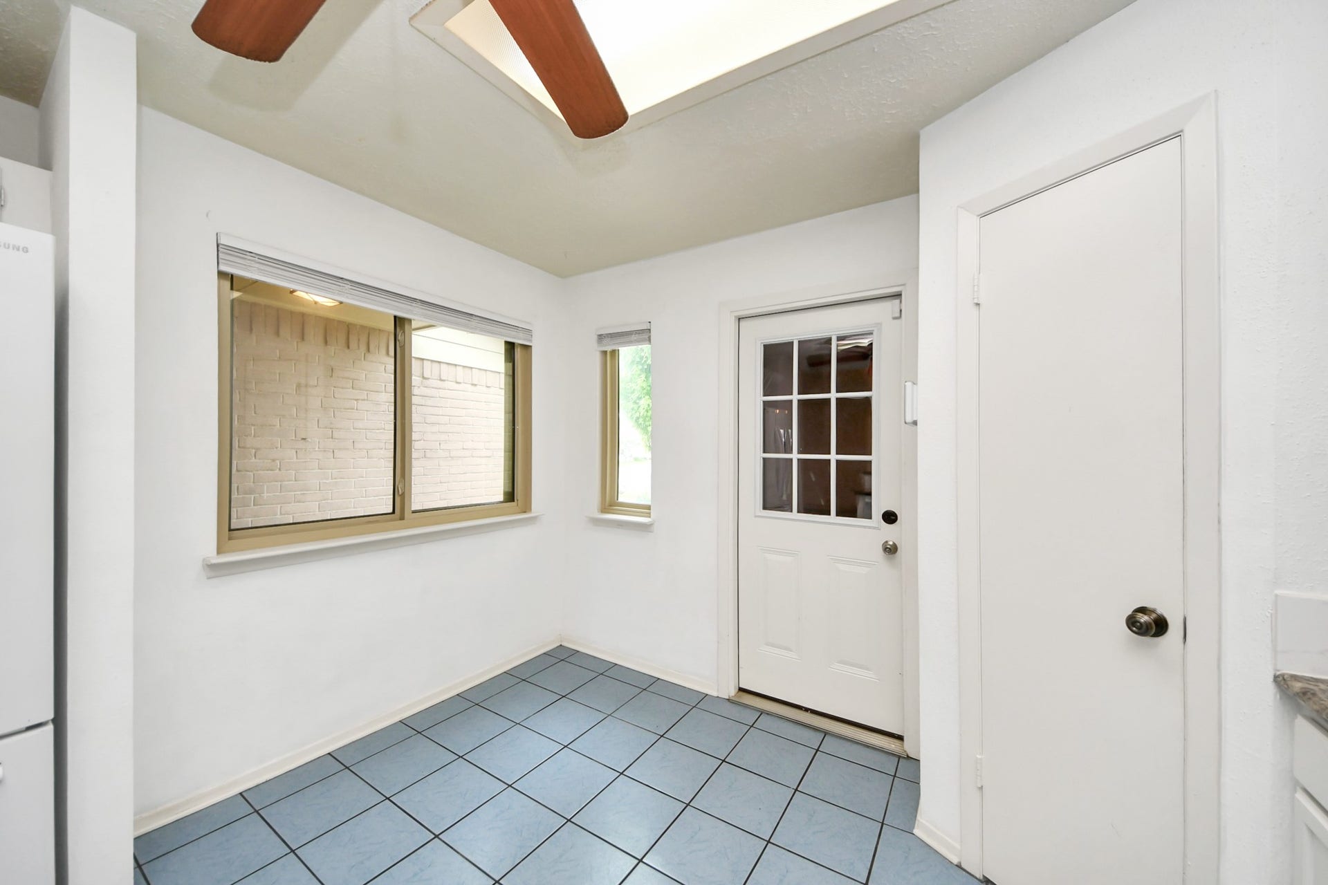 This bright room features a skylight, blue-tiled floor, and natural light from two windows. It includes a door with a glass panel leading outside and a white door to the side, offering a clean and versatile space.