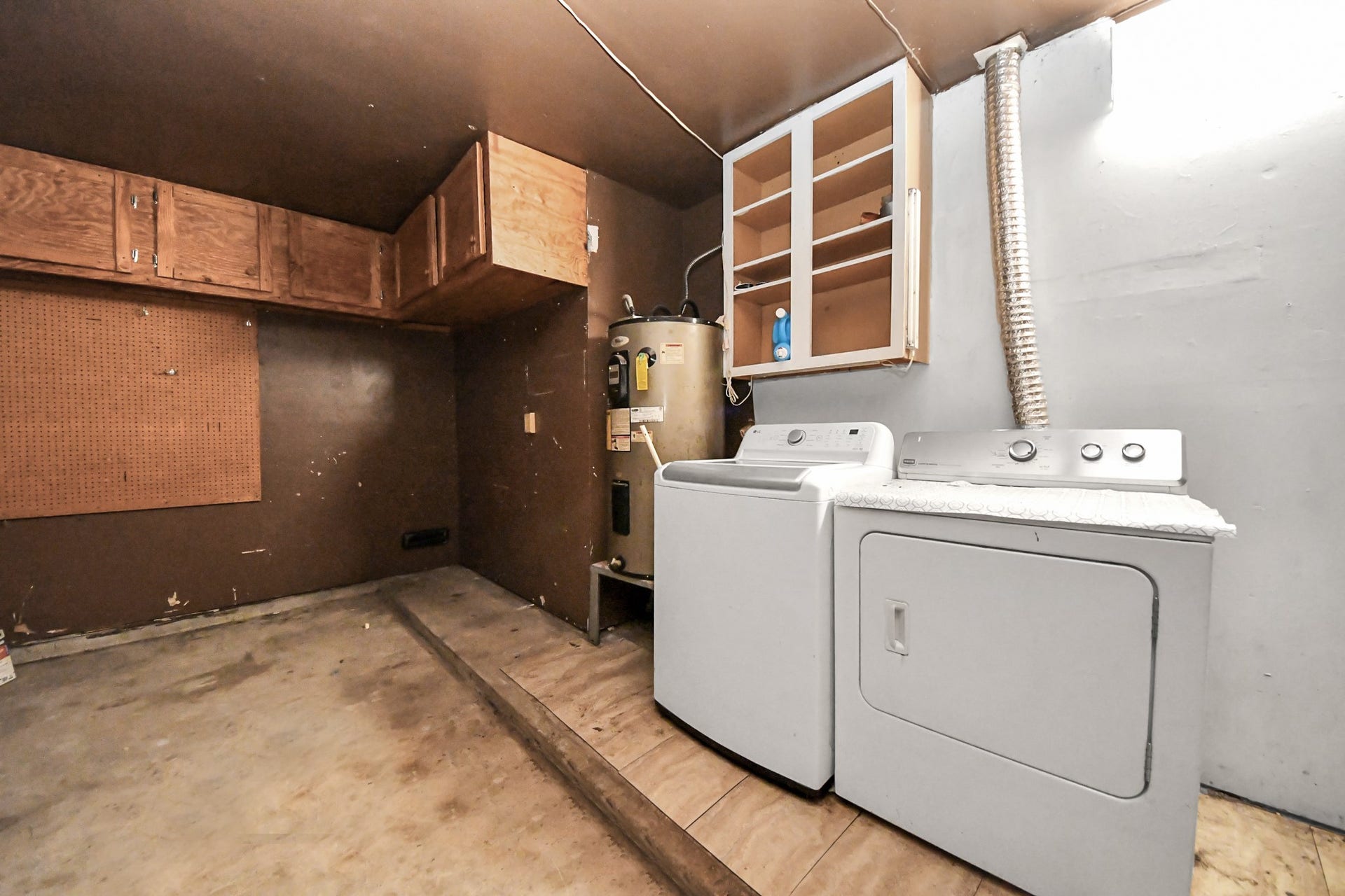 This photo shows a utility room with a washer and dryer, wooden storage cabinets, and a water heater. The space is functional with a pegboard for tools and a vent leading outside.