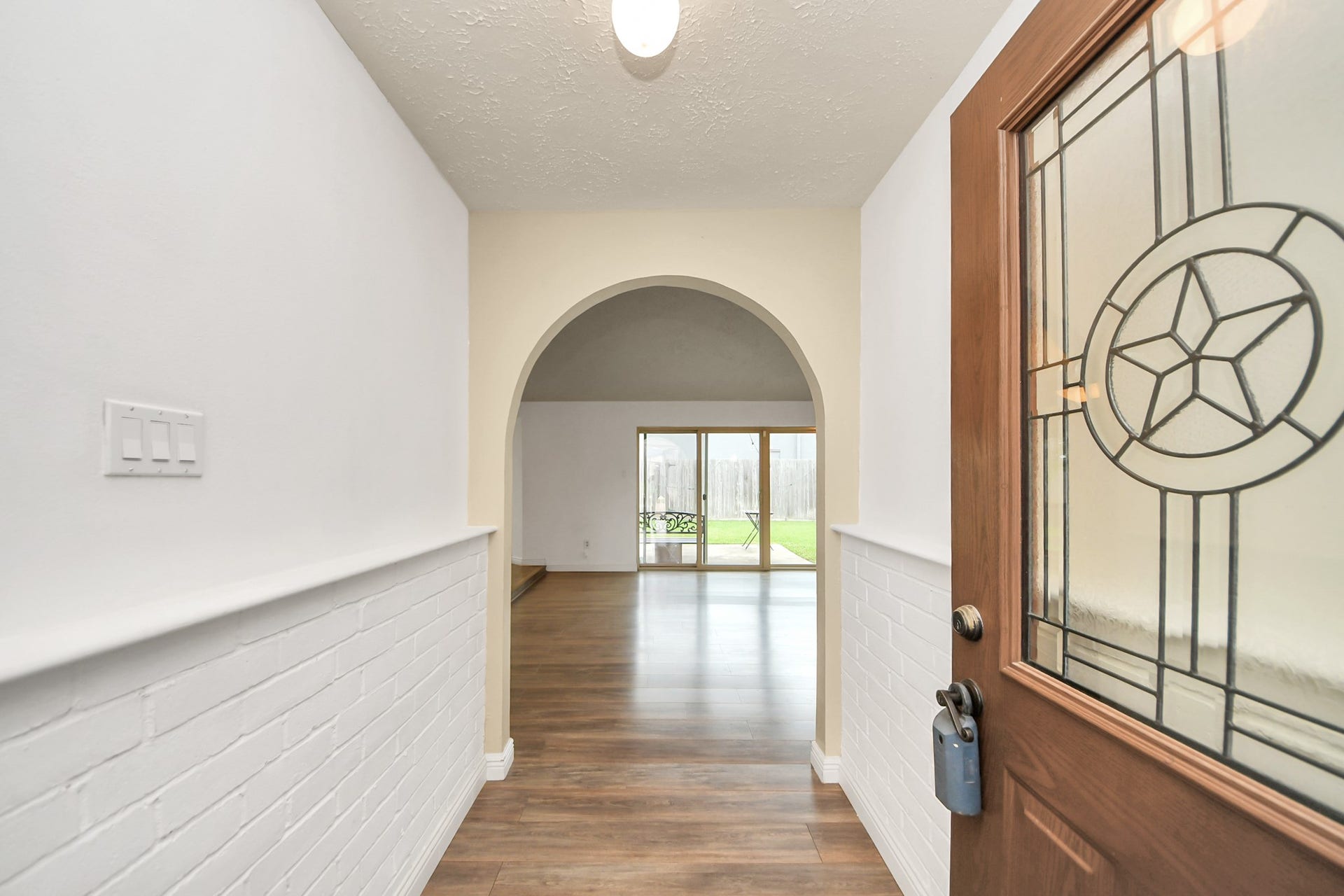 This entryway features a welcoming wooden door with decorative glass, opening to a bright hallway with white brick accents and hardwood flooring. An archway leads to a spacious room with large windows and a view of the backyard.
