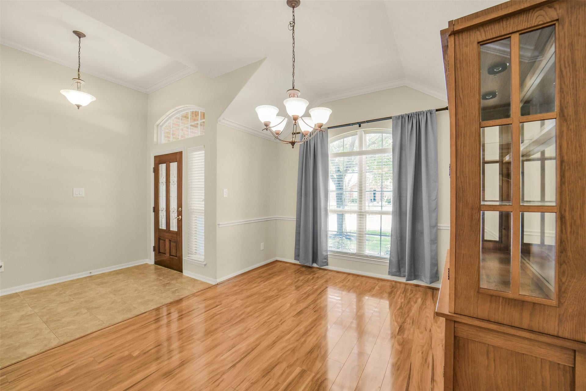 Elegant dining room featuring gleaming hardwood floors, a stylish chandelier, and abundant natural light from a large window with drapes.